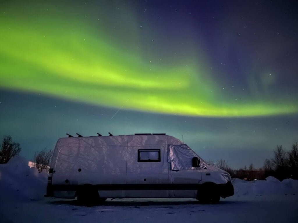 A campervan parked in the snow, under the green glow of the northern lights in Norway, on a winter vanlife trip. 