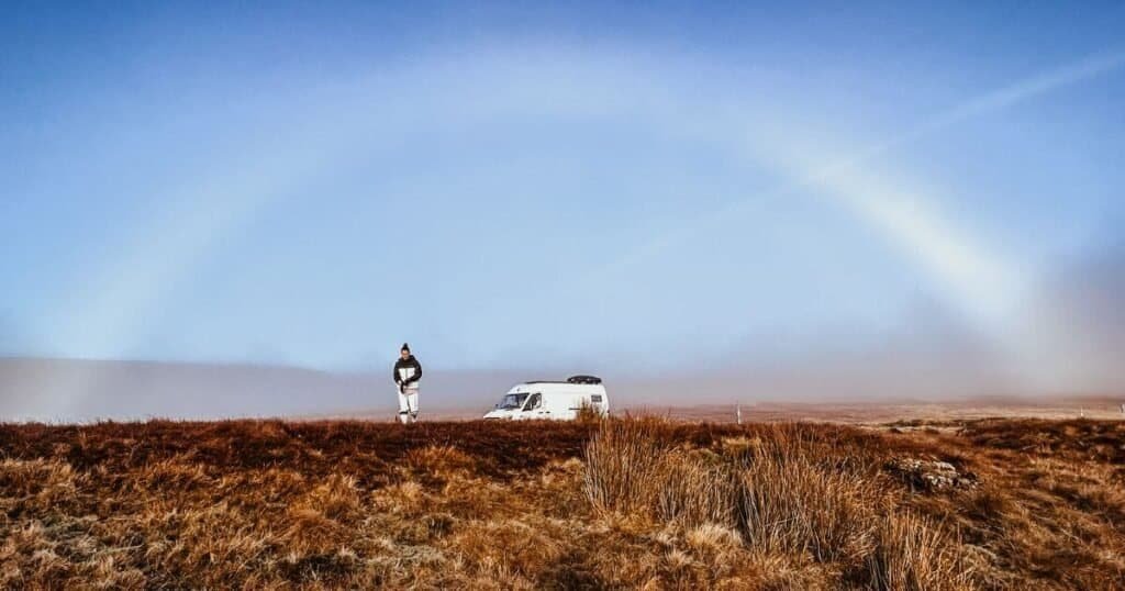 Woman walking away from a van life van, in the Yorkshire Moors amongst the heather, underneath a fogbow.