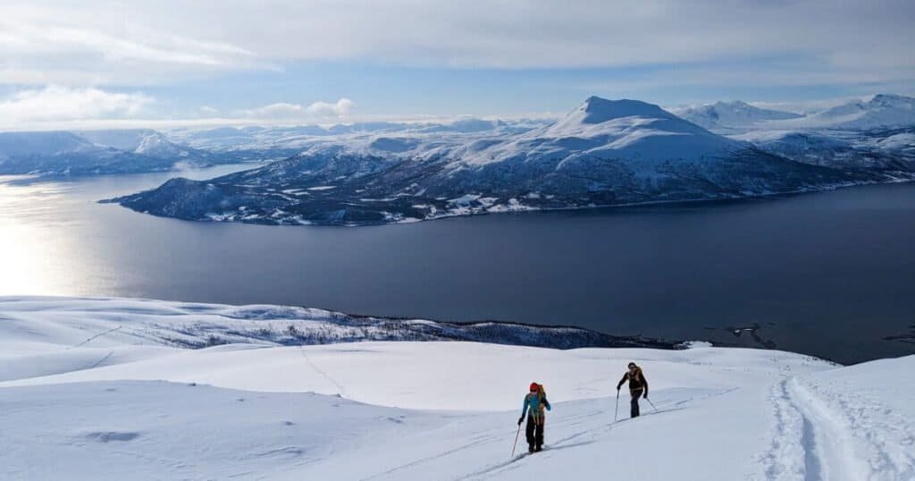 Two people ski touring off-piste on. a sunny day in norway. They are walking uphill in the foreground on a gentle snow covered slope and in the background there is a great lake and more snow covered peaks in the far distance