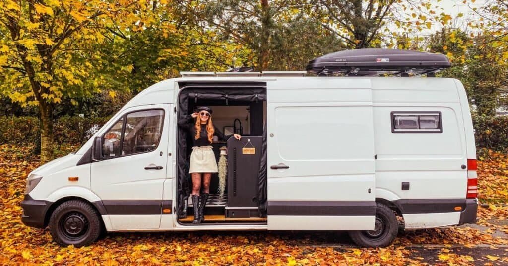 Woman stood in the side door of a converted campervan. Living van life in Ireland during Autumn, surrounded by Autumn leaves.