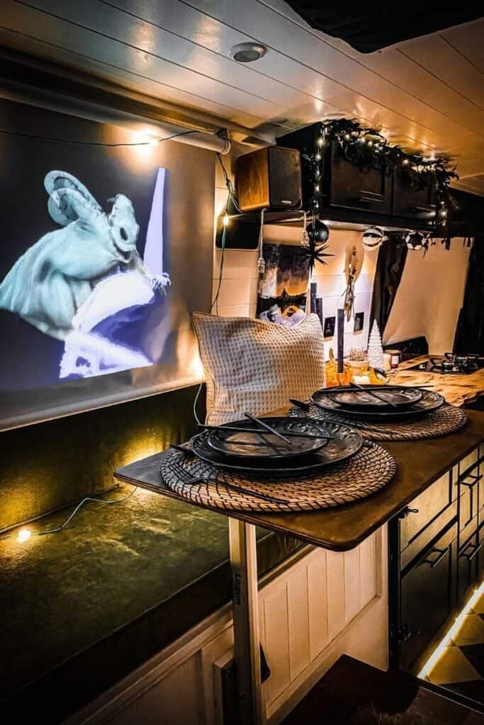 Inside of a campervan with gentle yellow mood lighting, white walls, black cabinets, and dark green sofa cushions. There is a black out blind pulled down with an image projected onto it.