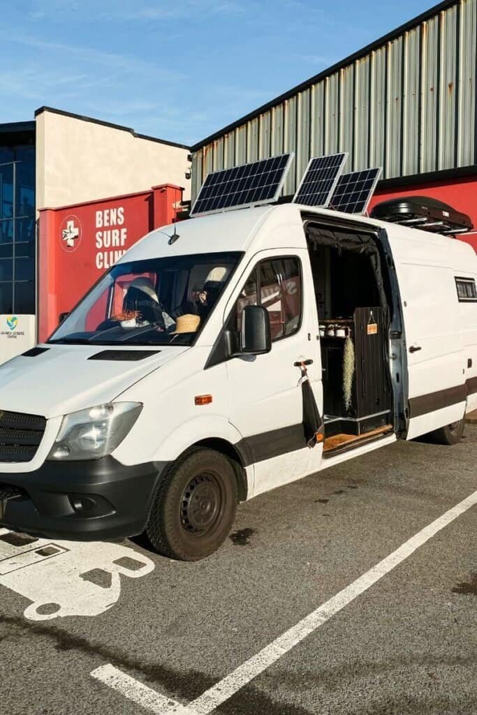 A white panel van parked on the tarmac with the side door open and the solar panels on the roof tilted upwards to get more solar. Very useful during winter vanlife.