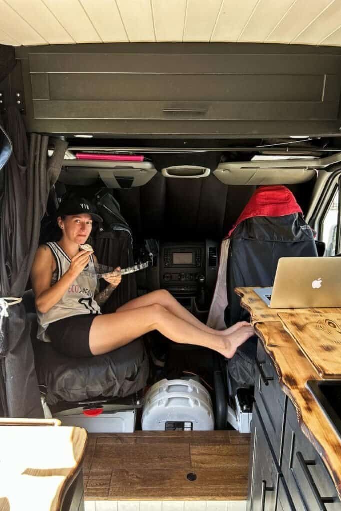 A woman sat in the front of a van with her legs across both front chairs and the chairs are spun 180 degrees to face the back