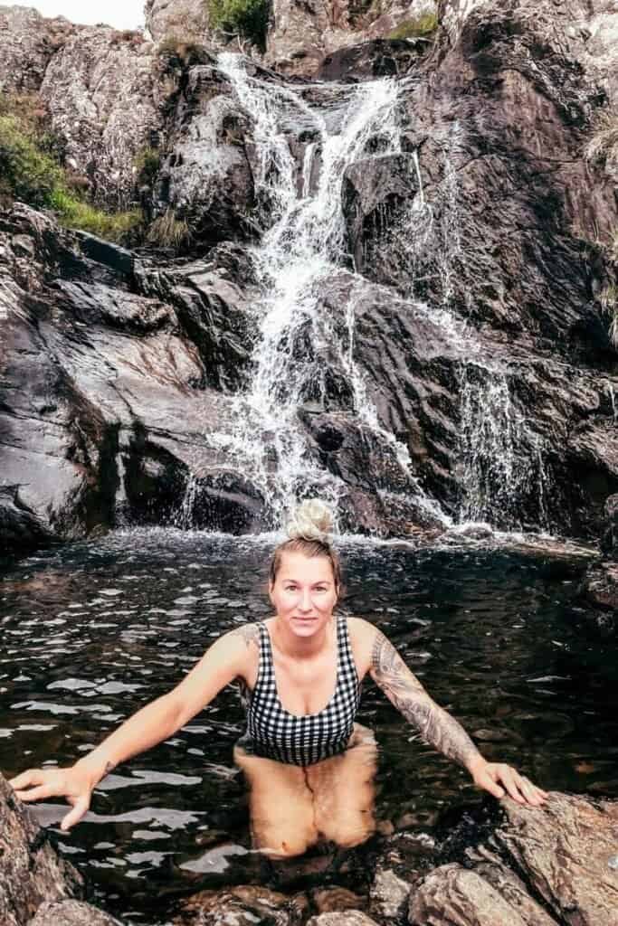 Woman in black and white checkered swimsuit, squatted in a small rock pool as a small waterfall cascades behind her. Ben Hope, Scotland. Nature bathing is an alternative option for vanlifers. 