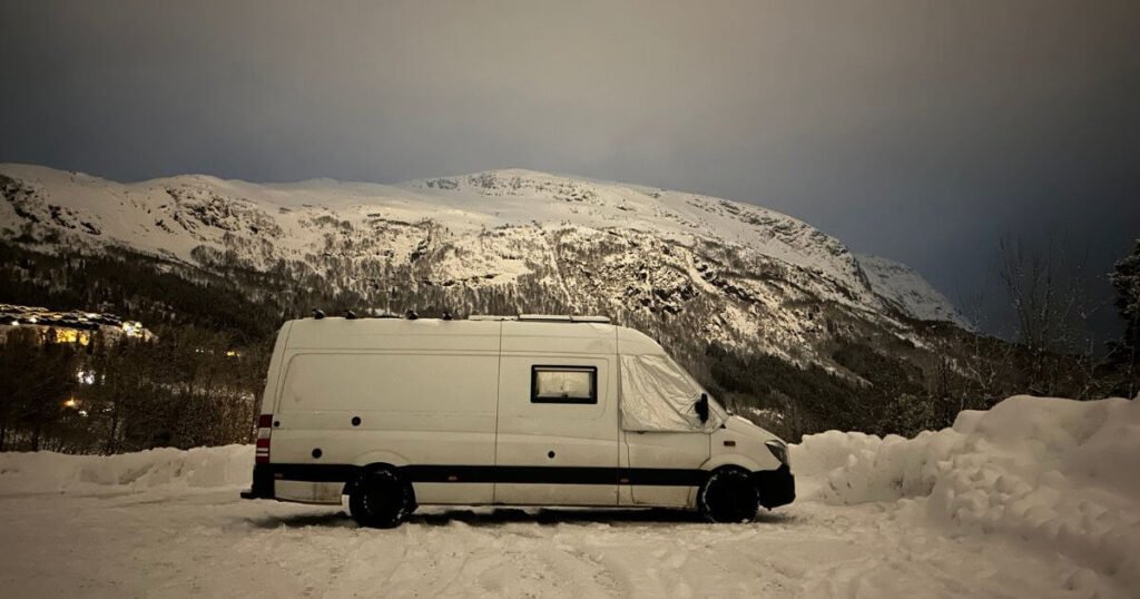 Side view of a mercedes sprinter van with. asnow covered mountain in the background. The van has a silver insulation cover wrapped around the outside of the front windows, to help battle against the extreme winter vanlife cold. 