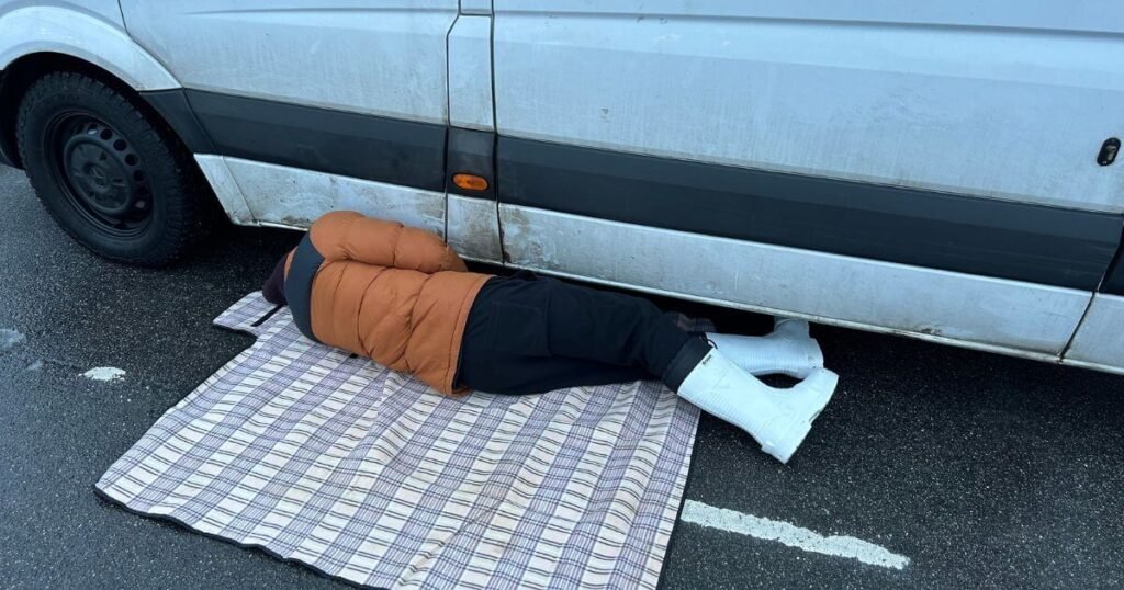 A person in an orange jacket, from behind, lying on their side on tarmac peaking inderneath a white van. 