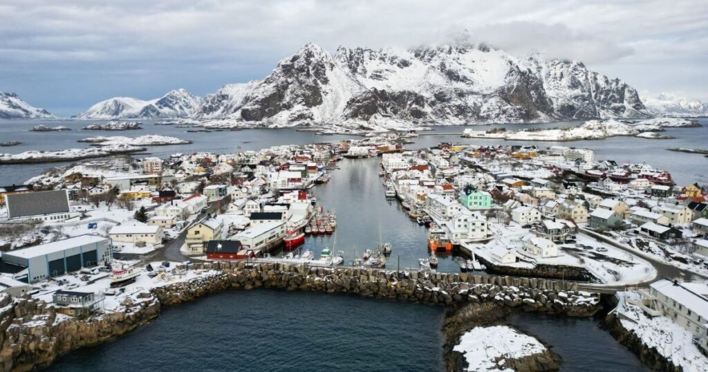 A drone shot looking out on a snowy tiny village in norway covered in snow, based slightly out to sea. The village is surrounded by water and roads linking small areas of land to the mainland in the background which has giant white mountain peaks as far as the eye can see.