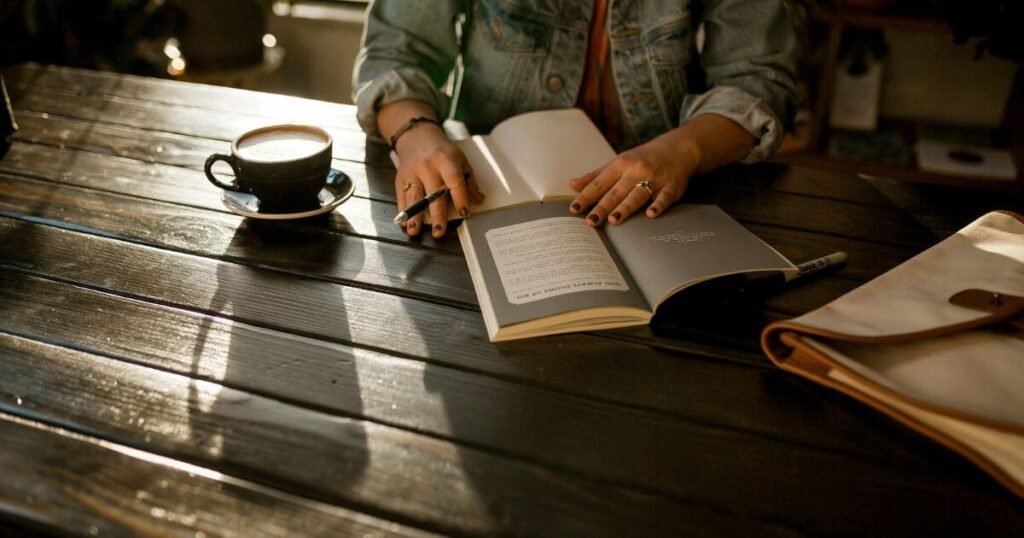 Woman sat at a table with a coffee, taking notes on a book.