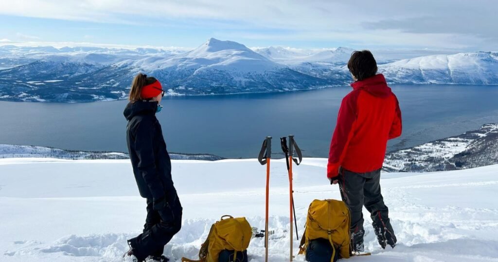 Two people stood in the snow on the side of a mountain, looking out at a large lake and snow covered peaks in the distance, during a winter vanlife adventure.