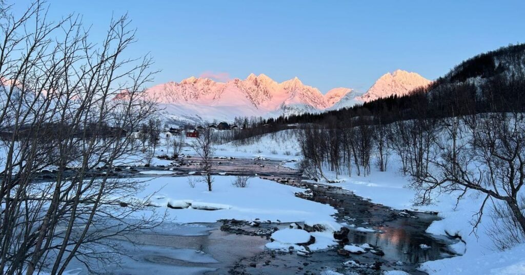 A frozen river surrounded by snowy banks in the foreground with peach coloured sunlit snowy mountain peaks in the background.