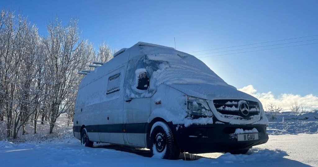 A front on view of a Mercedes Sprinter van covered in, and surrounded by snow during a winter vanlife roadtrip. There is bright blue sky and snow covered trees in the background.