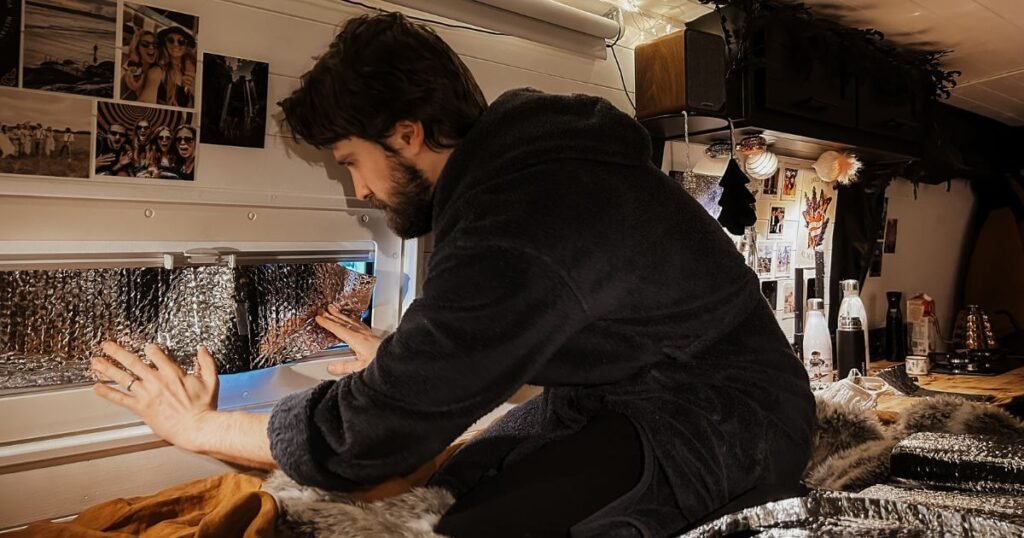 A man in a dressing gown in bed, in a campervan interior, adding insualtion foil to his interior window to help plug a little more of that winter vanlife chill. 