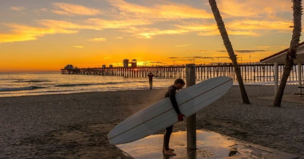 Surfer holding surfboard while showering at the beach at sunset.