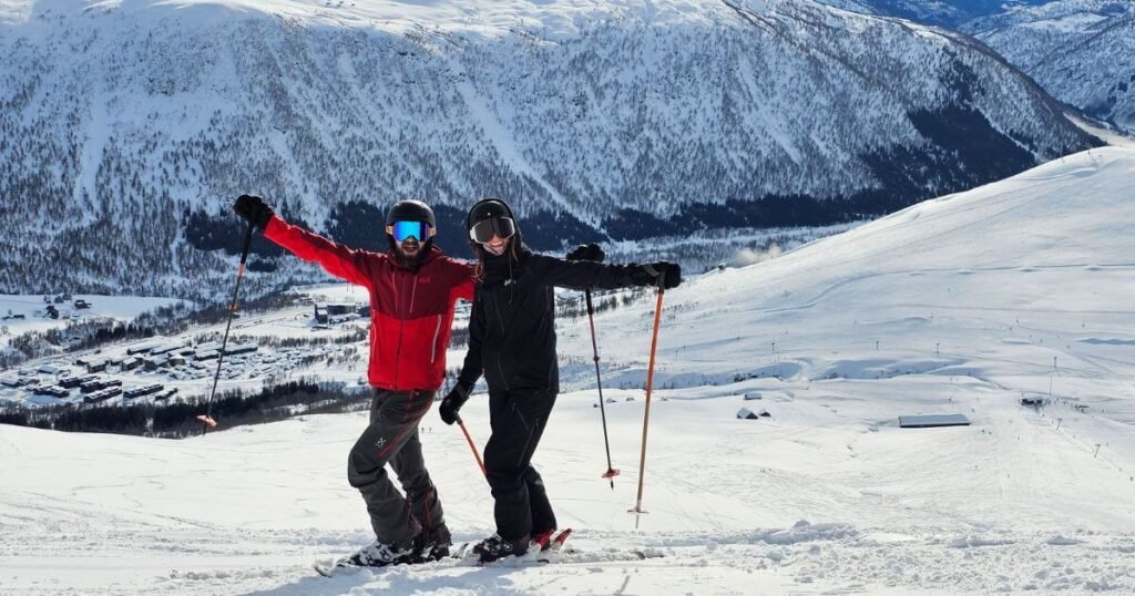 Two people in ski gear waving at the camera. In the background is a snow covered ski slope and a snow covered valley with tree dotted in the distance.