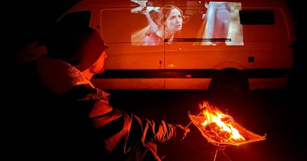 A man sat beside a small fire pit watching a movie projected onto the side of a white van during a cold night in winter vanlife.