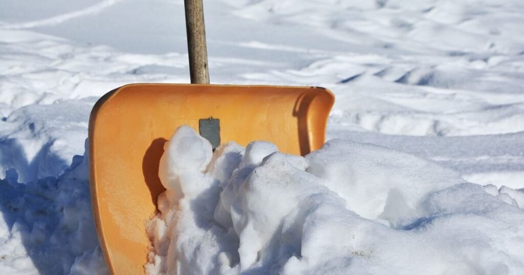 A close up of an orange snow shovel handle cutting through snow on the ground getting ready to clear it. During a day in winter vanlife. 