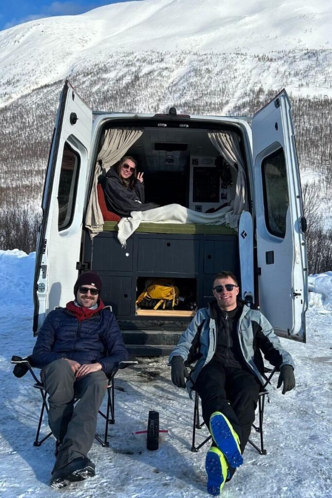 Looking at the back of a large panel van during winter vanlife. The doors are swung open and a women is smiling and making a peace sign at the camera from a bed in the back. In front of the van are two men sat in chairs smiling at the camera. 