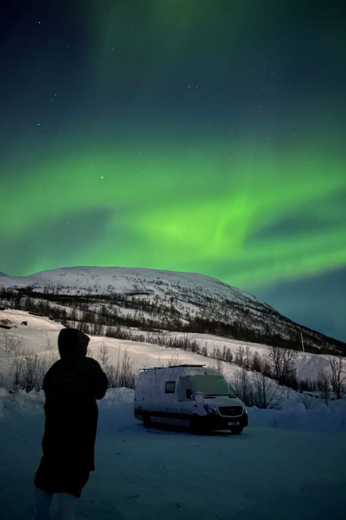 A nighttime winter vanlife setting. A van parked in the snow, within snowy mountain peaks behind it and green aurora lighting up the sky above. 