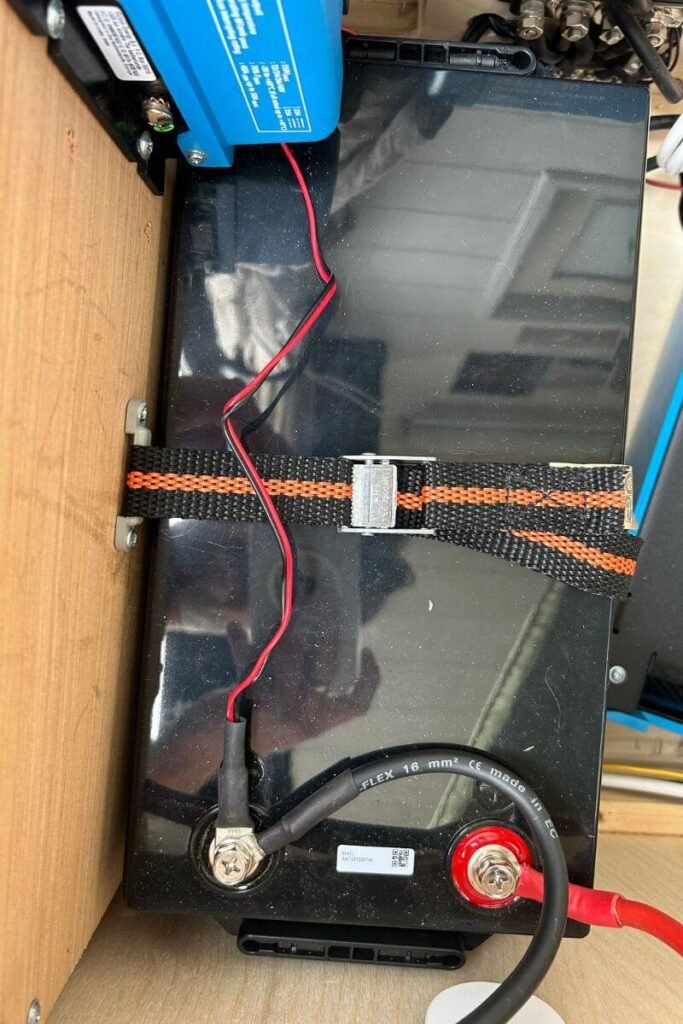 Looking down on the top of a big black leisure battery in a wooden compartment. Black and red wires can be seen connecting to it. 