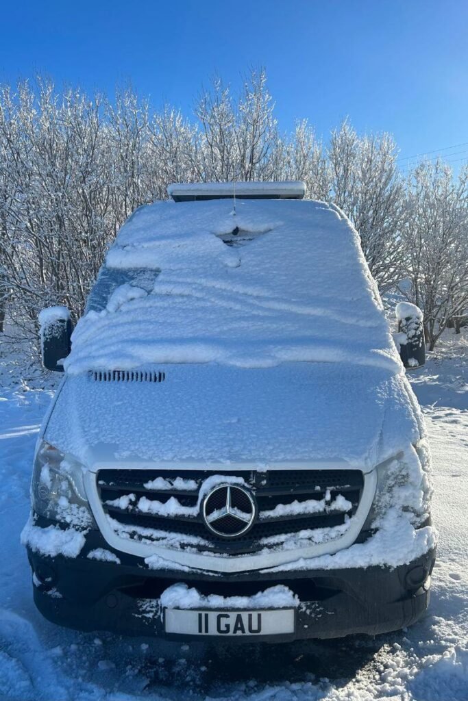 Close up of the front of a van covered in a few inches of snow during winter vanlife. There is blue sky in the background. 