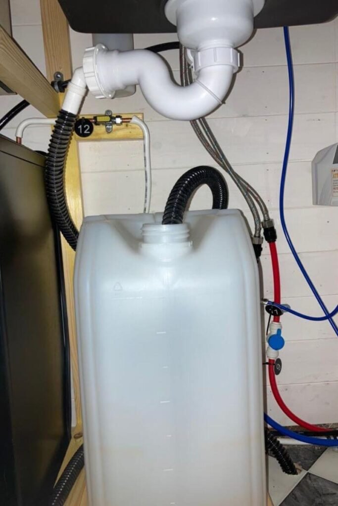 A 20 Litre grey water tank with black piping going into the top, positioned underneath the white u-bend of a sink. This is an alternative plumbing option for winter vanlife to keep all water inside the heated vehicle. 