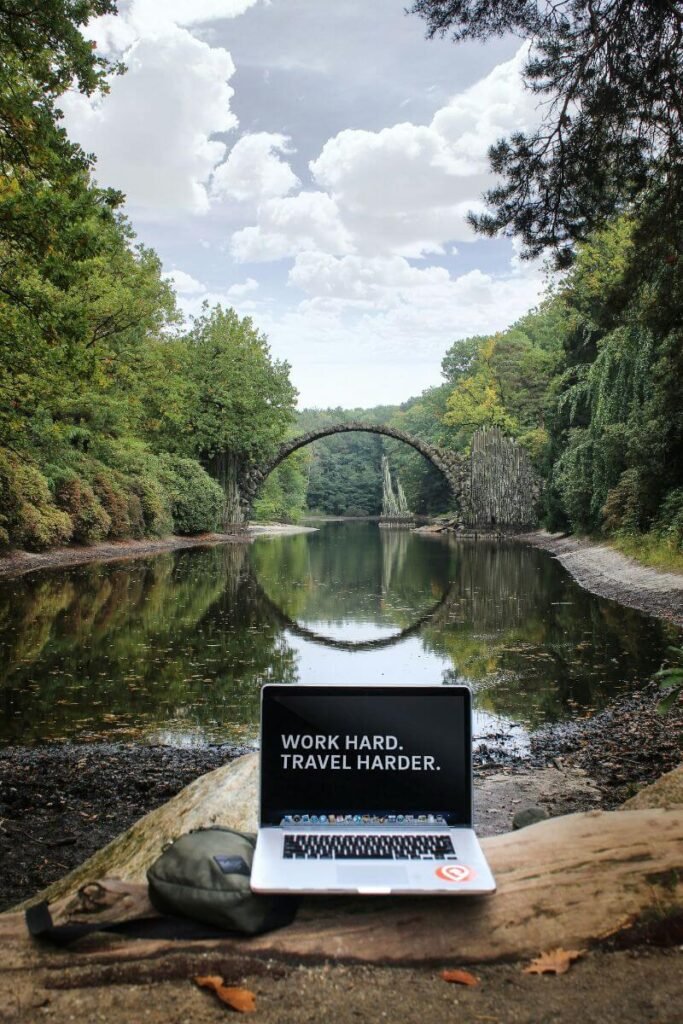 Lake, trees and a bridge with a circular reflection. In front is a laptop, the screen says "work hard, travel harder".