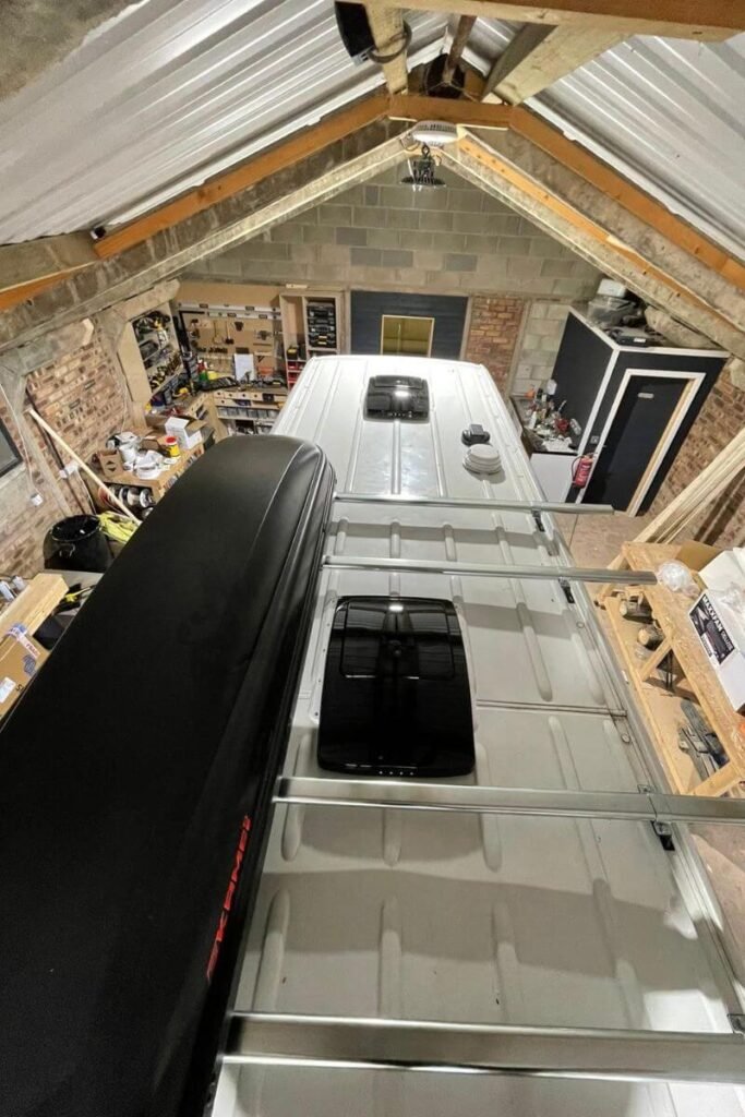 A view of the top of a white van inside a workshop. On the roof there are two black square roof vent fans, one at the front and one at the back. 