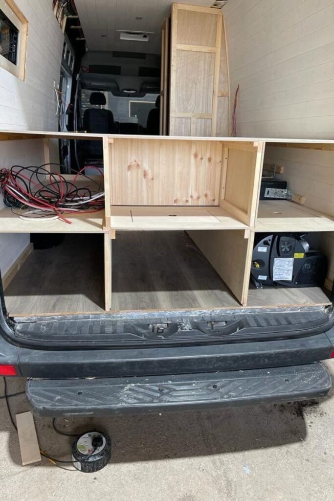 A view inside the back door of a van, with white wooden cladded walls. There is a brown wooden framework structure erected in the back housing electrical wiring in the top left and a black boiler in its own compartment in the bottom right. 