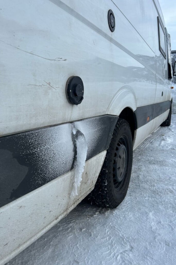 Close up on the side of a white van. Below a black circular exhaust vent there is a frozen cascade of water, due to extreme temperatures during a winter vanlife trip. 