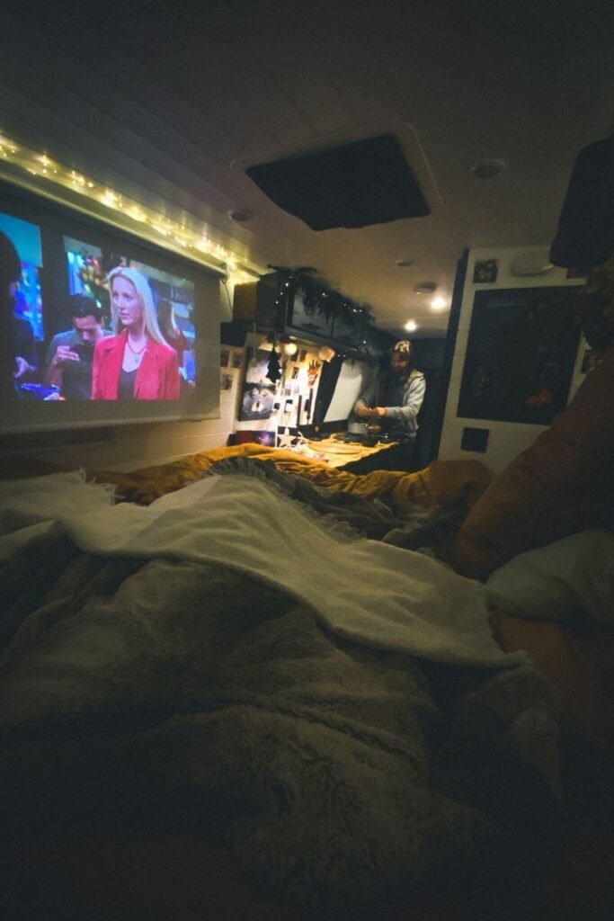 The cosy inside of a campervan, in dim light. The bed is covered in lots of blankets and a projector is projecting a the friends TV show onto a black out blind. A cosy night in winter vanlife. 