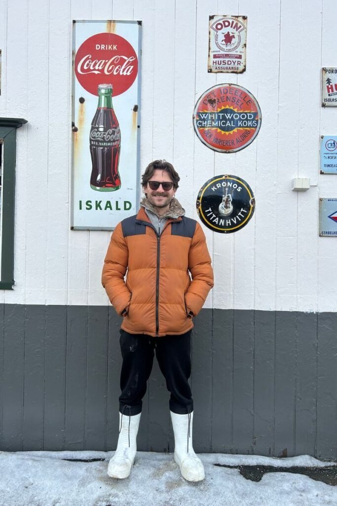 A man in an orange winter coat, wearing big white winter boots stood smiling at the camera in front of a white wall. 