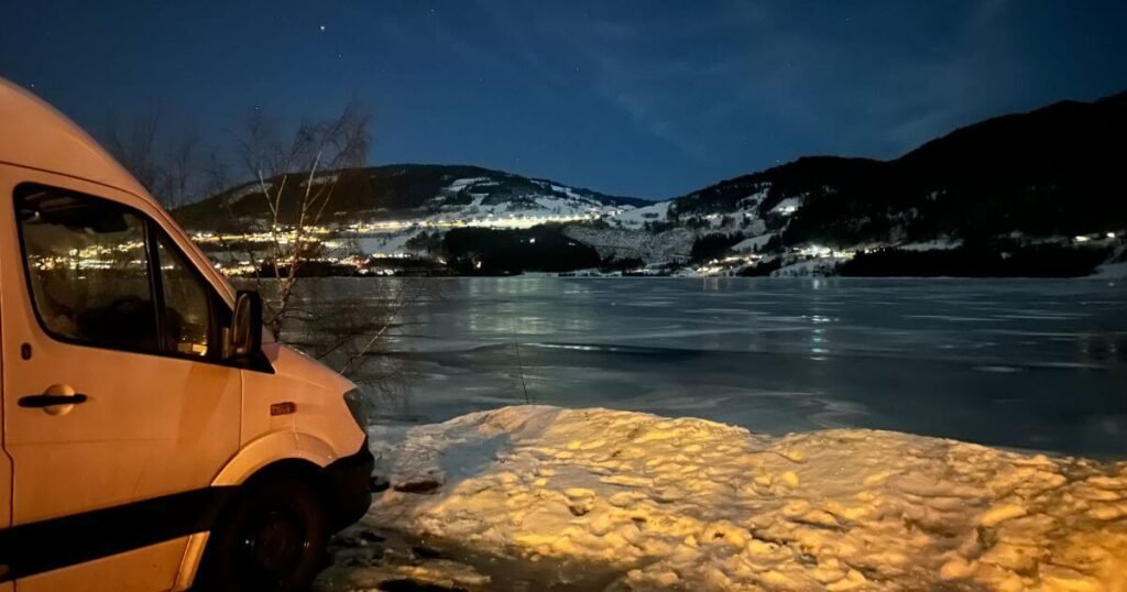 A white van parked by a frozen lake at night time with snow cover mountains in the background during a cold winter.